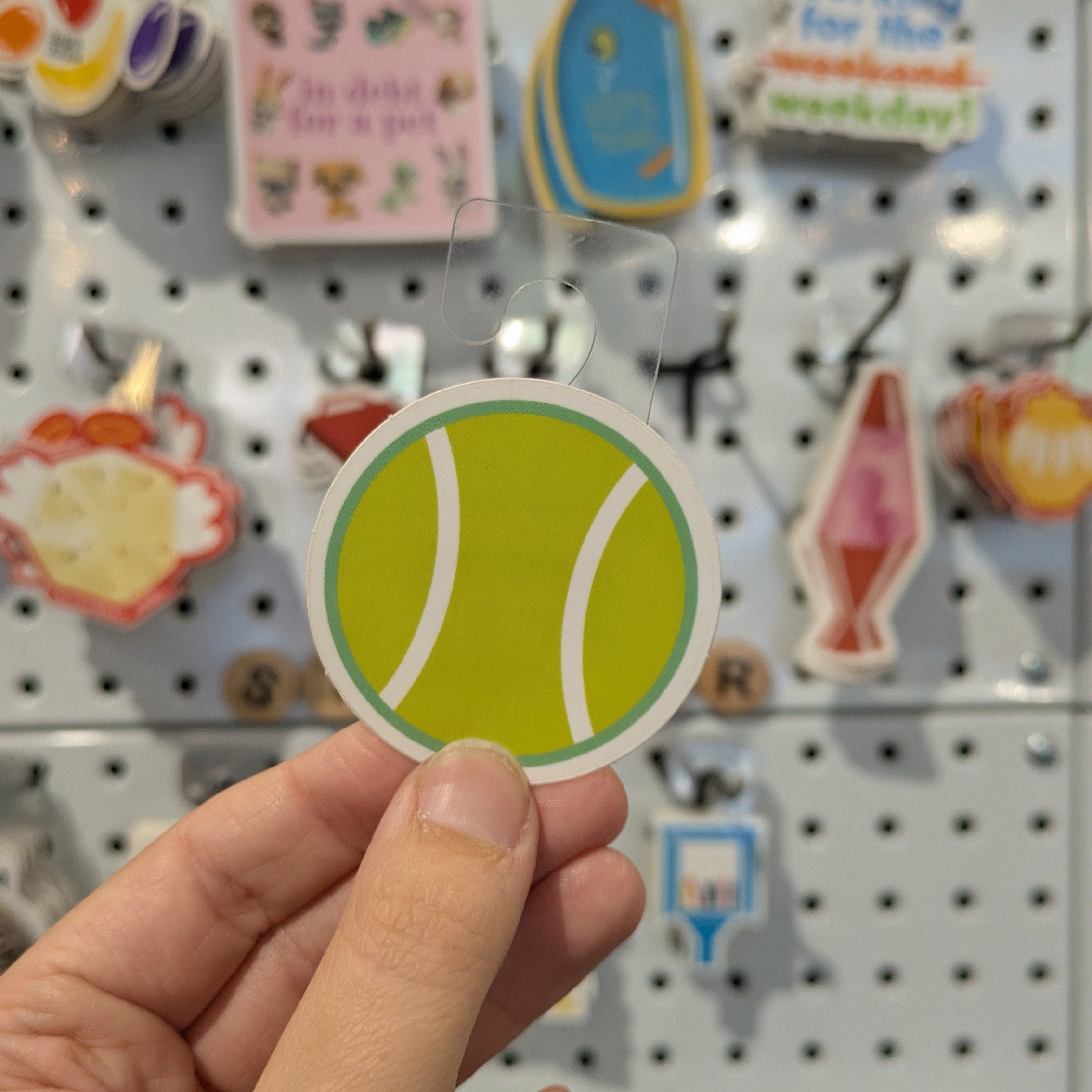 Hand holding a round green sticker with a white circle, against a pegboard background with various other stickers.