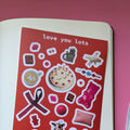 A sticker sheet with various designs including a teddy bear, hearts, and other romantic-themed elements, with a red background and the text 'love you lots'.