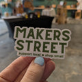 Hand holding a 'MAKERS STREET' sticker with a blurred background of a store interior.