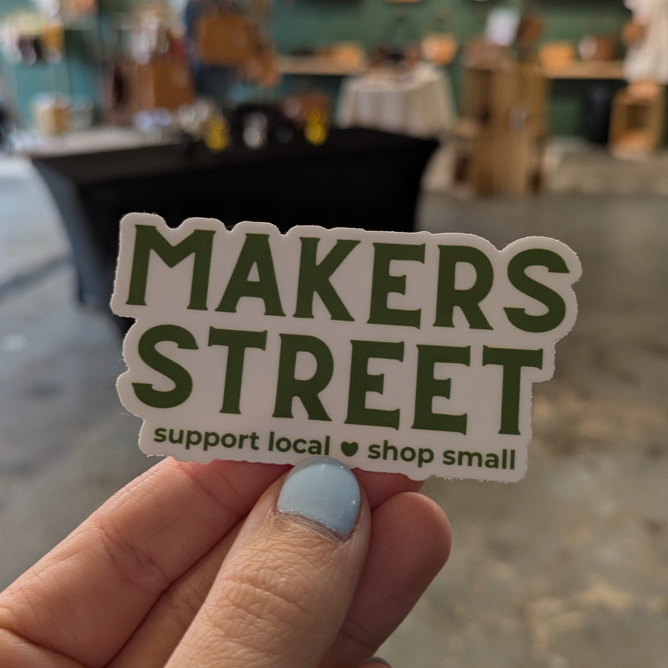 Hand holding a 'MAKERS STREET' sticker with a blurred background of a store interior.