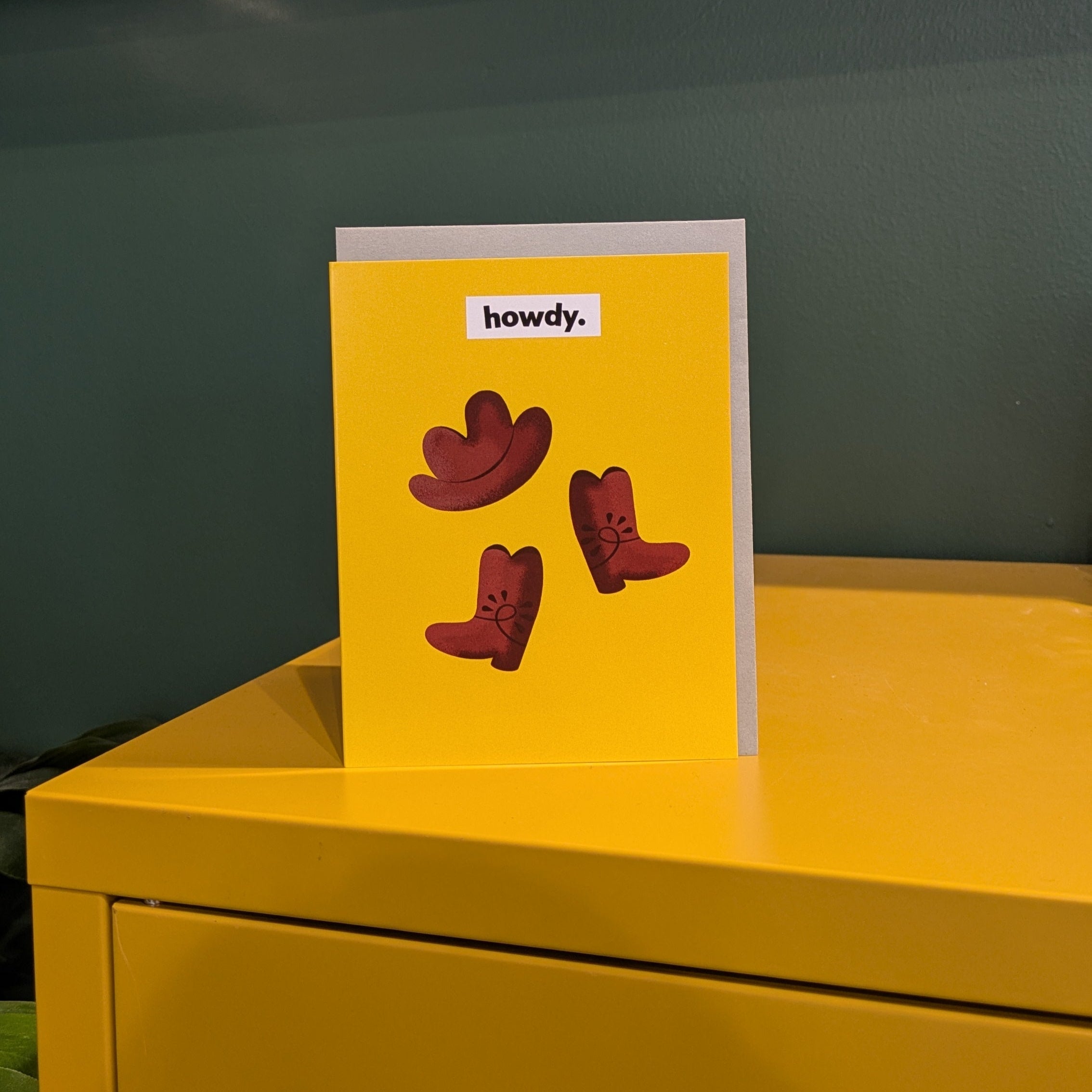 Yellow card with an  illustrated cowboy hat and boots on a yellow surface against a green wall.