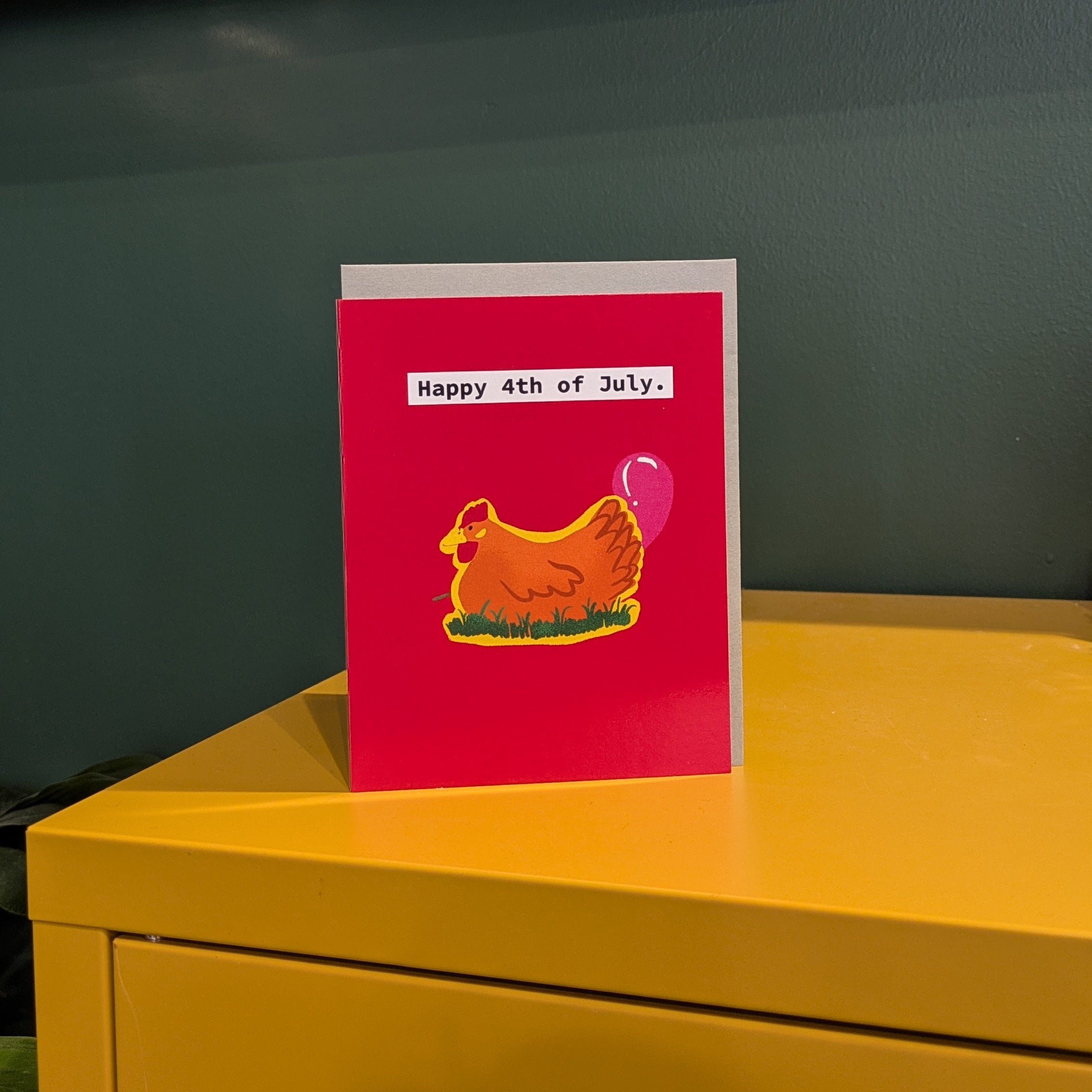 4th of July Card with a red background and a yellow chicken design on a yellow surface.