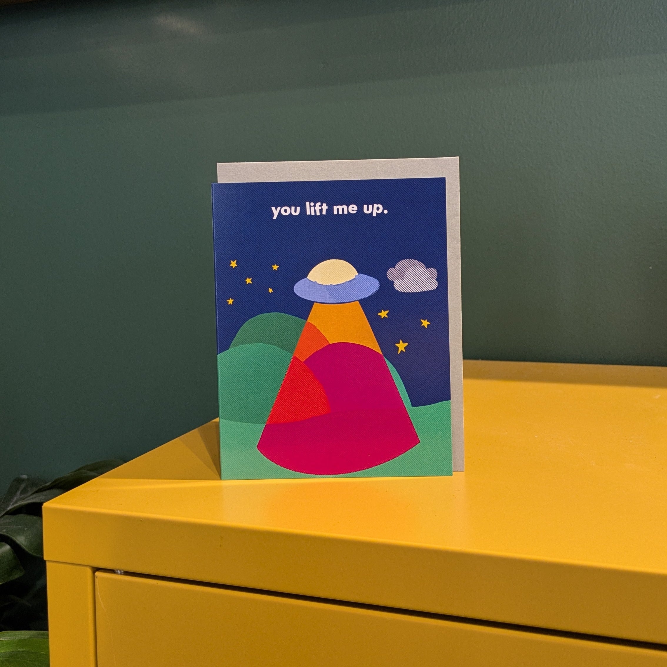Colorful greeting card "you lift me up" on a yellow surface with more books on a shelf in the background.