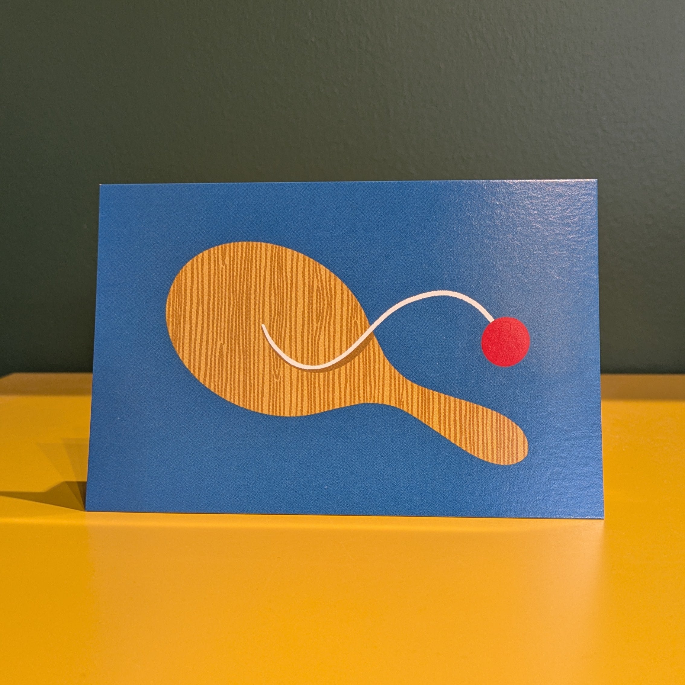 Card with a wooden paddle and red ball on a blue background, placed on a yellow surface.