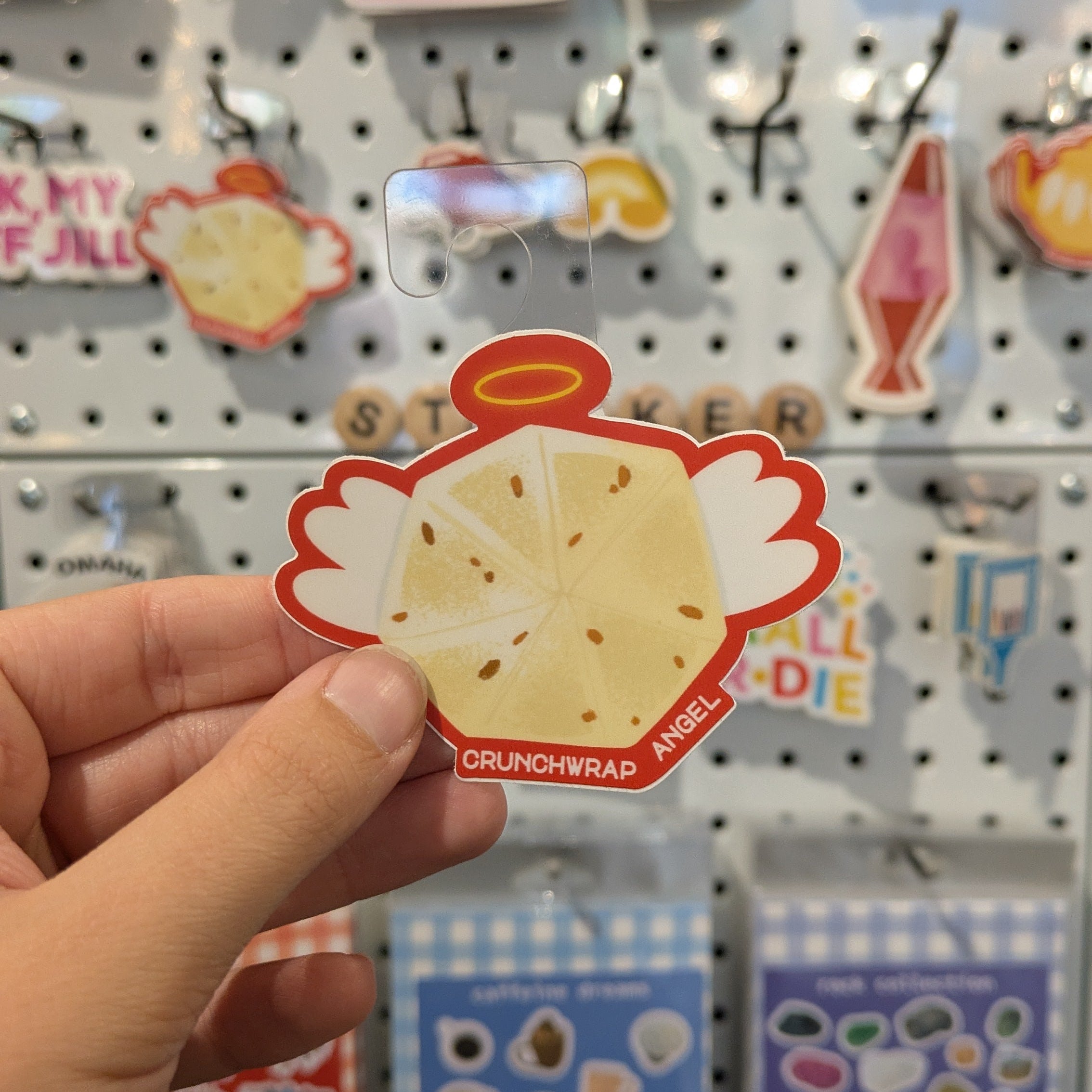 Hand holding a crunchwrap sticker with a character on a pegboard with various other stickers.