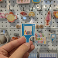 Hand holding a small blue and white little library sticker with text against a background of various colorful signs on a pegboard.