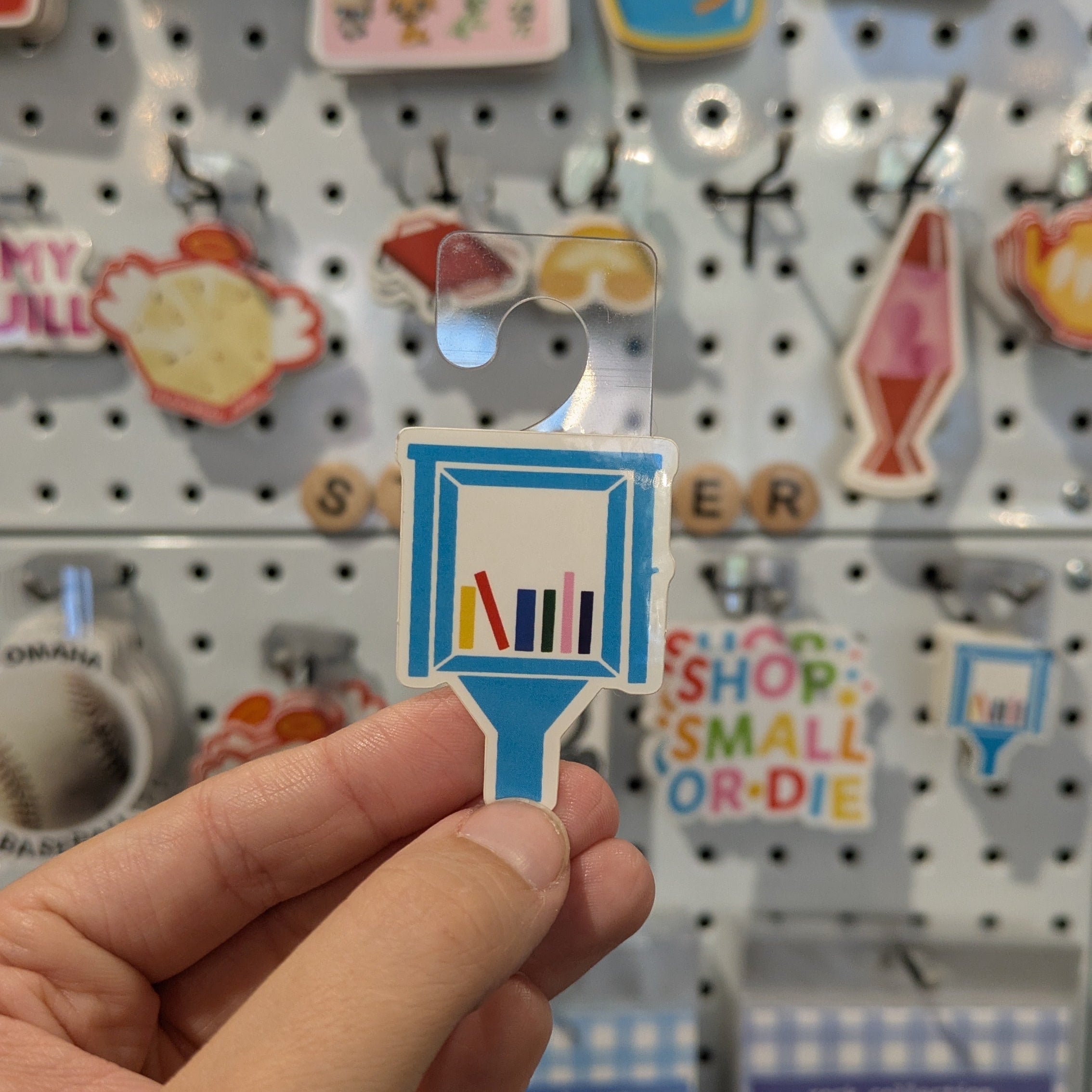 Hand holding a small blue and white little library sticker with text against a background of various colorful signs on a pegboard.