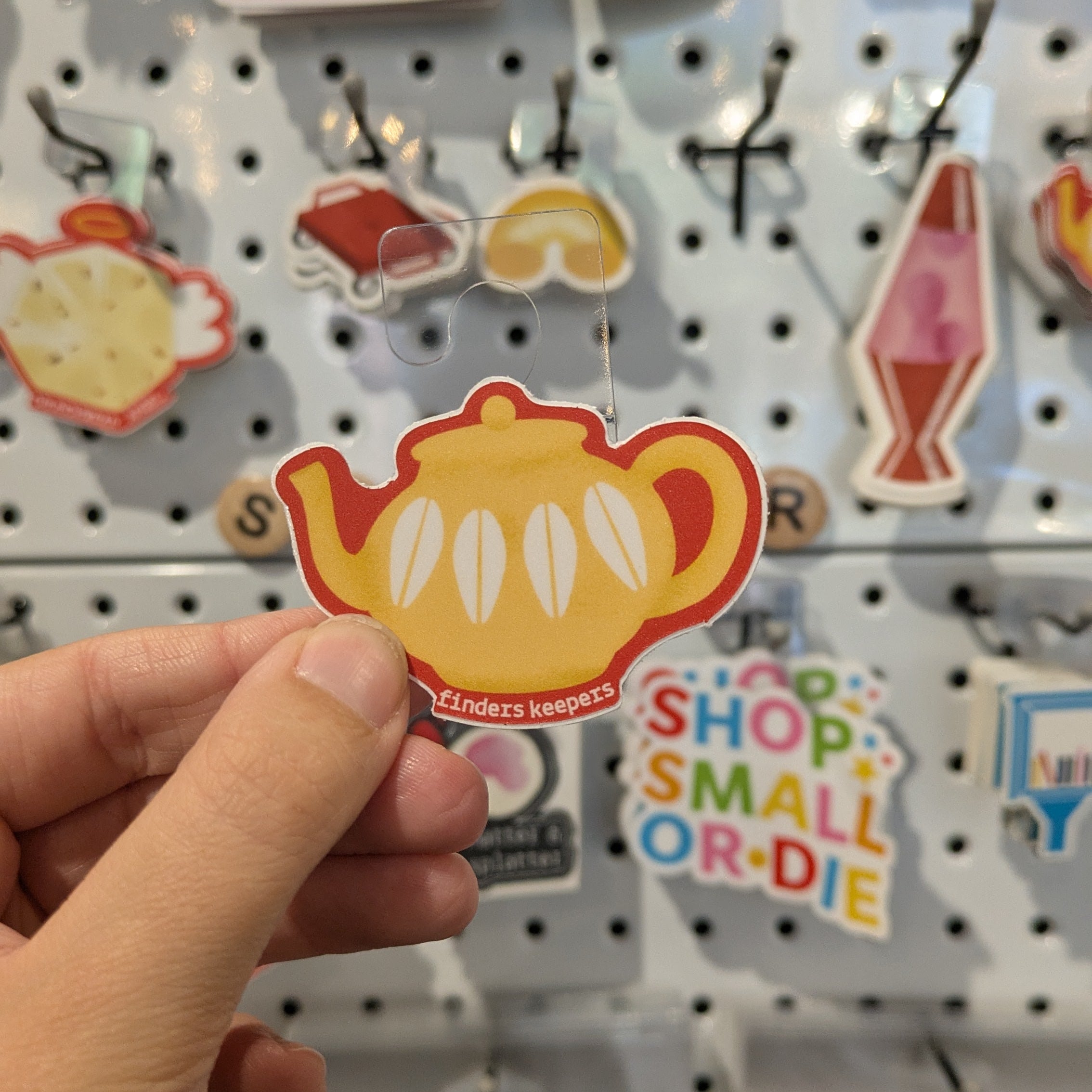 Hand holding a decorative sticker with a teacup design against a pegboard background with various other stickers.