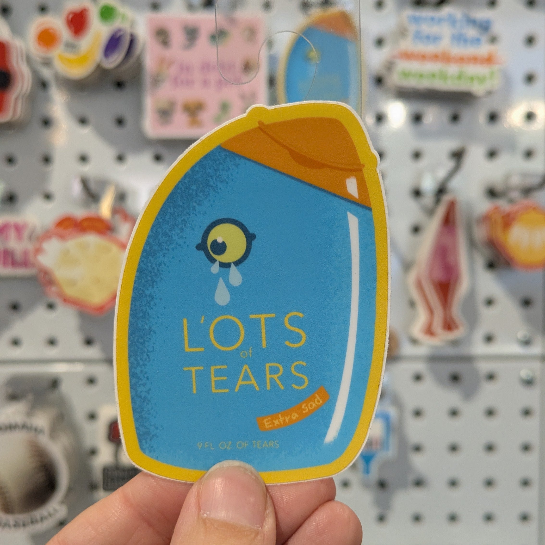 Sticker with 'LOTS TEARS' text held by a hand against a pegboard background with various other stickers.