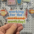 Hand holding a colorful magnet with 'working for the weekend weekday!' text against a pegboard background.