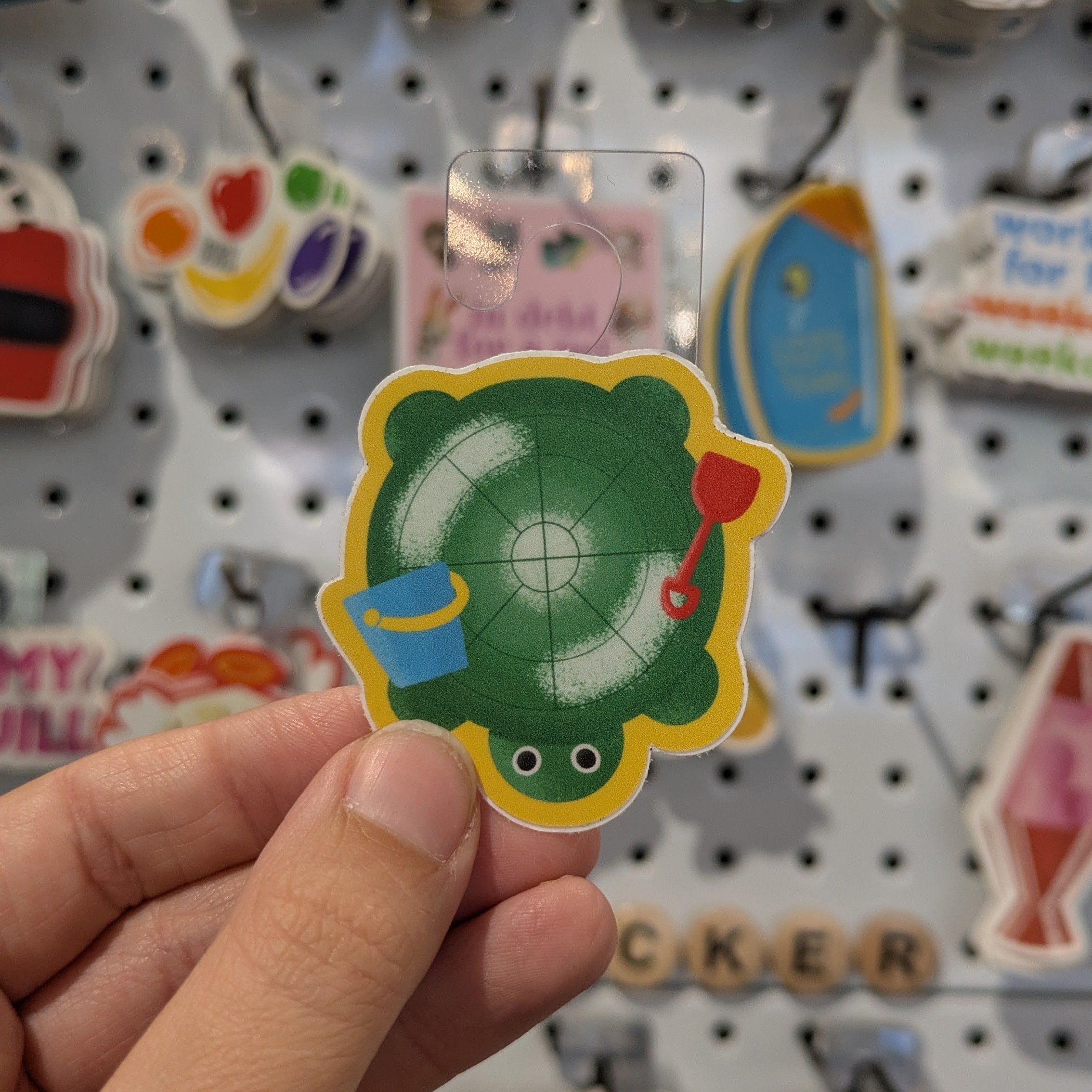Hand holding a colorful sticker with a turtle design against a pegboard background.
