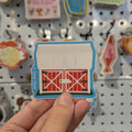 Hand holding a sticker of a red barn against a pegboard with various other stickers.