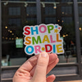 Hand holding a colorful 'Shop Small or Die' sticker in front of a building with large windows.