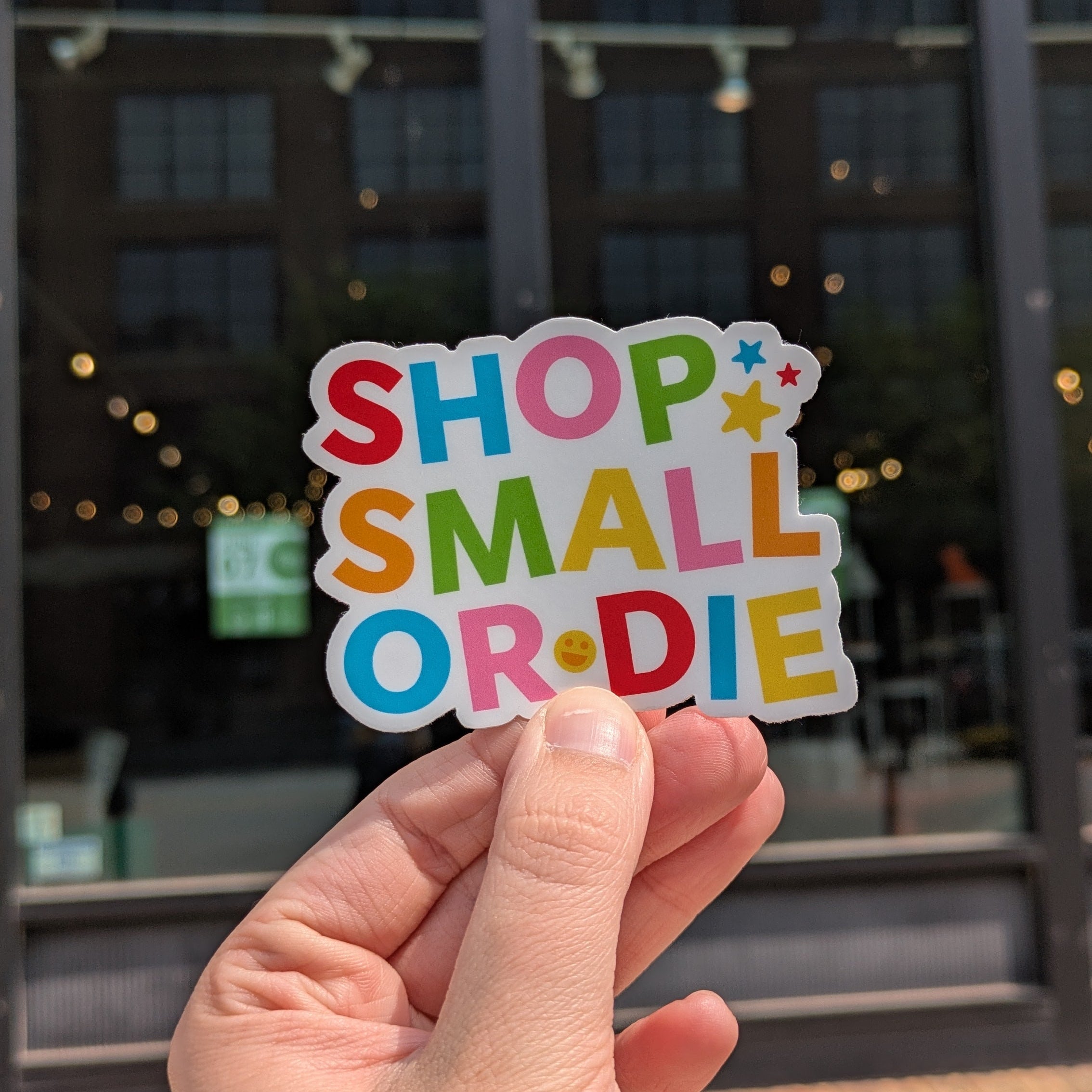 Hand holding a colorful 'Shop Small or Die' sticker in front of a building with large windows.
