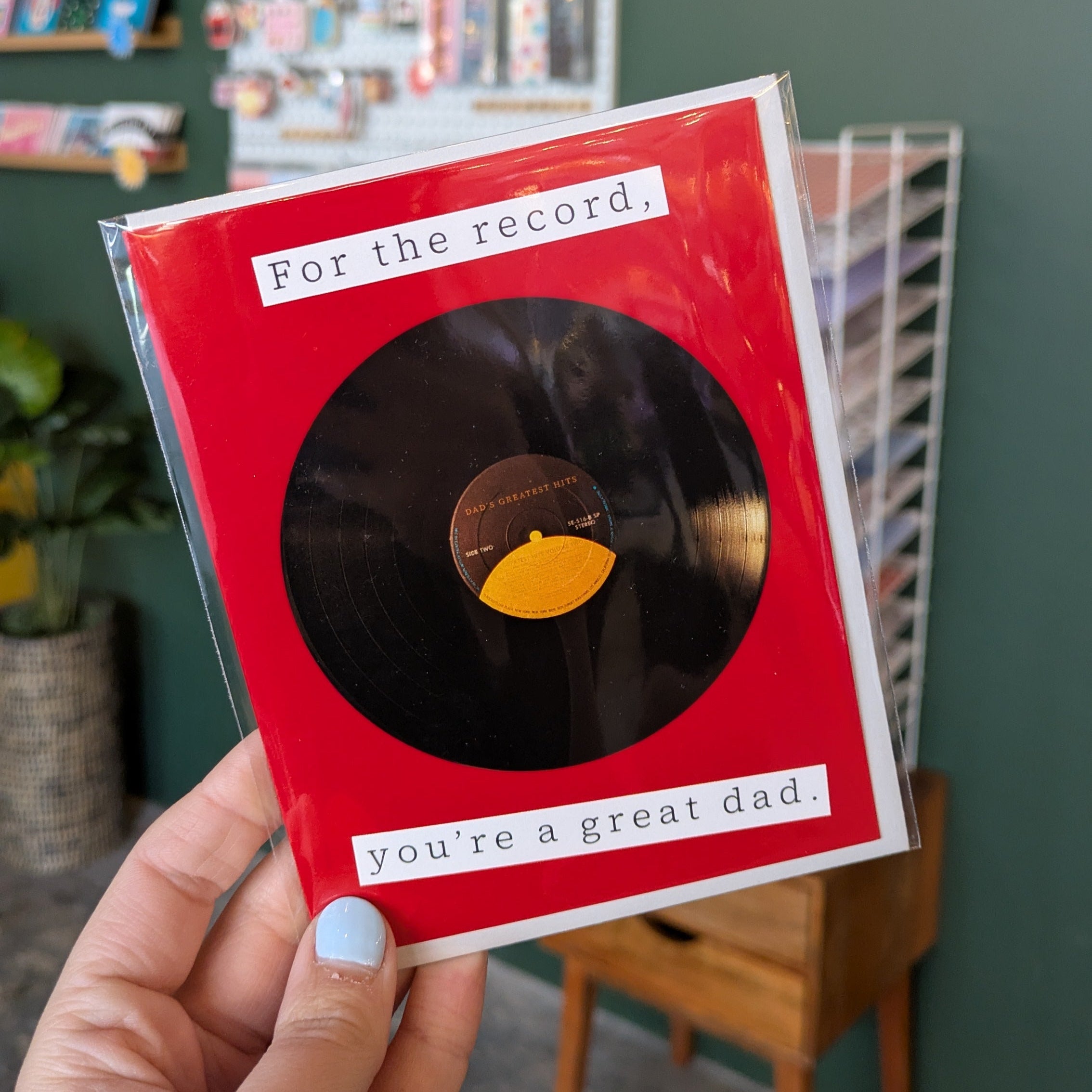 Hand holding a red card with a vinyl record design and text in a casual indoor setting.