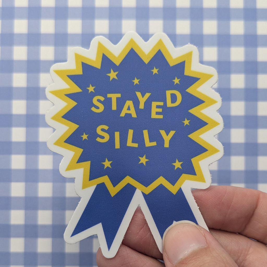 A hand holding a die-cut sticker shaped like a blue ribbon award badge with yellow stars and the text 'STAYED SILLY' in yellow, set against a blue and white checkered background.