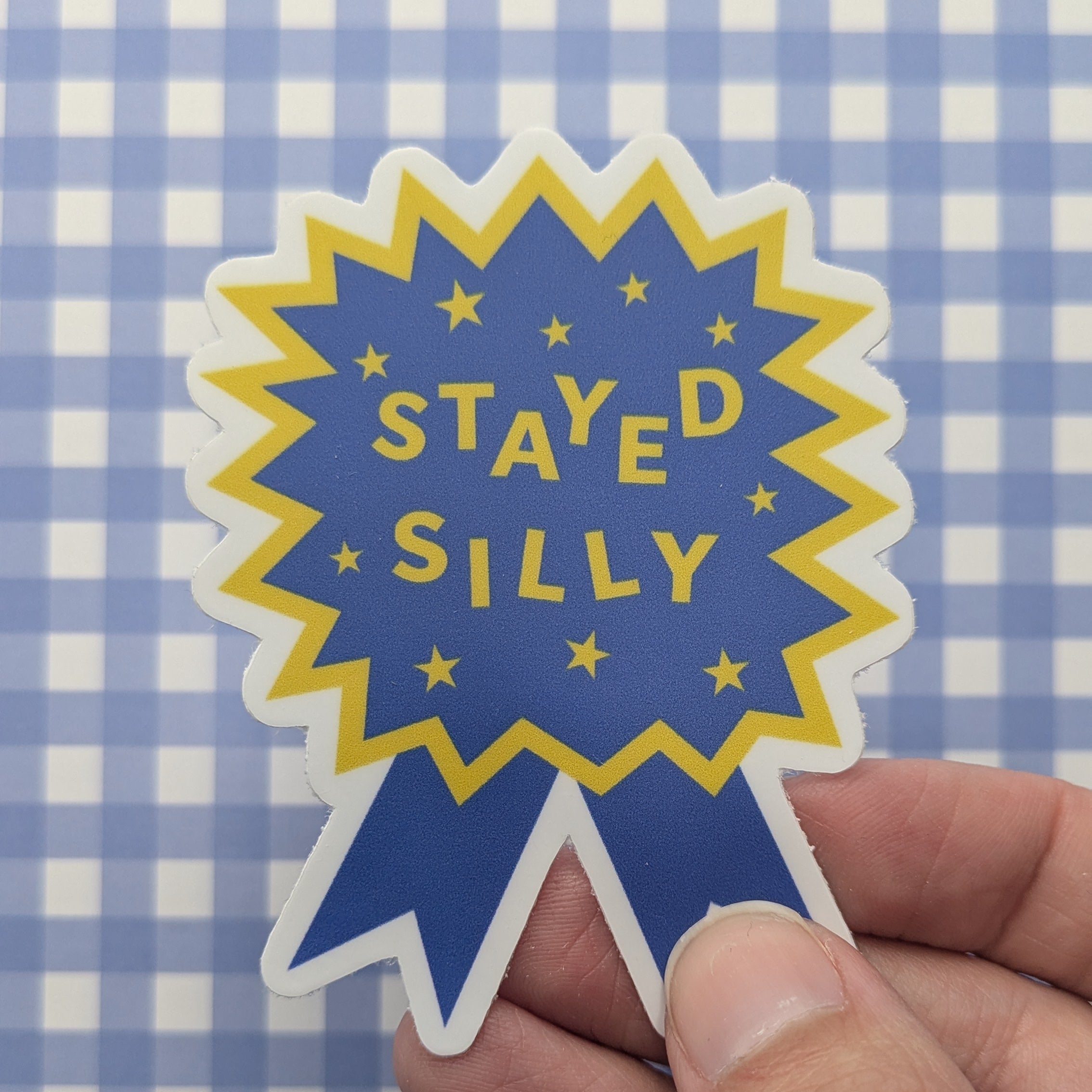 A hand holding a die-cut sticker shaped like a blue ribbon award badge with yellow stars and the text 'STAYED SILLY' in yellow, set against a blue and white checkered background.