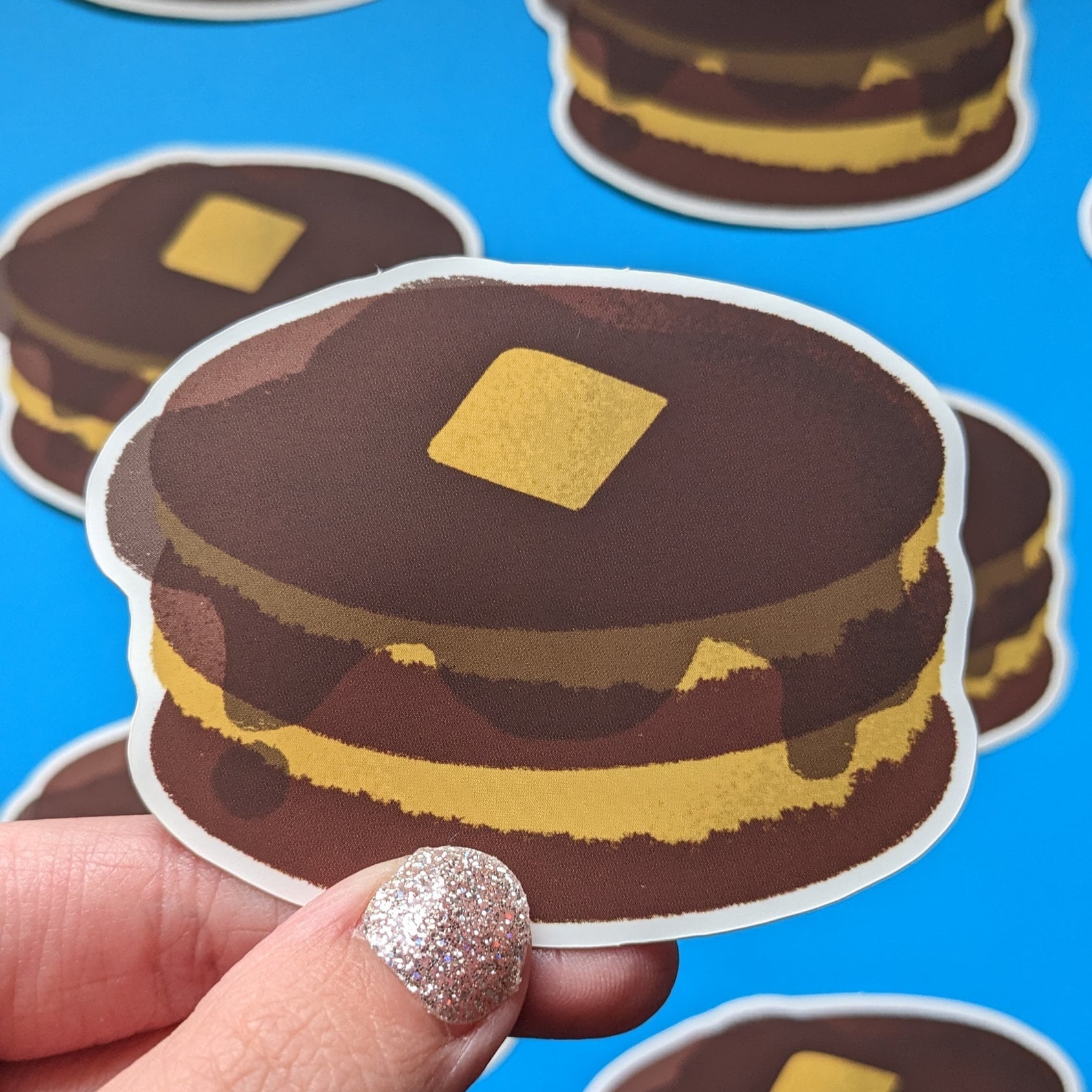 A sticker with a design of a stacked pancake on a blue background, held by a hand with a glittery manicure.
